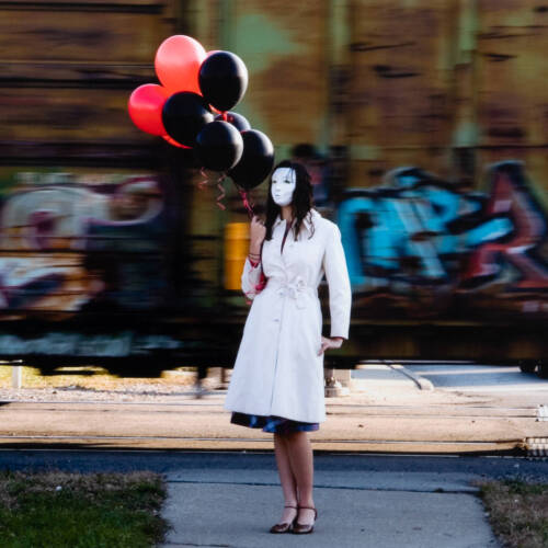 Color photograph of a person in a white coat, a white mask covering their face, holding red and black balloons.